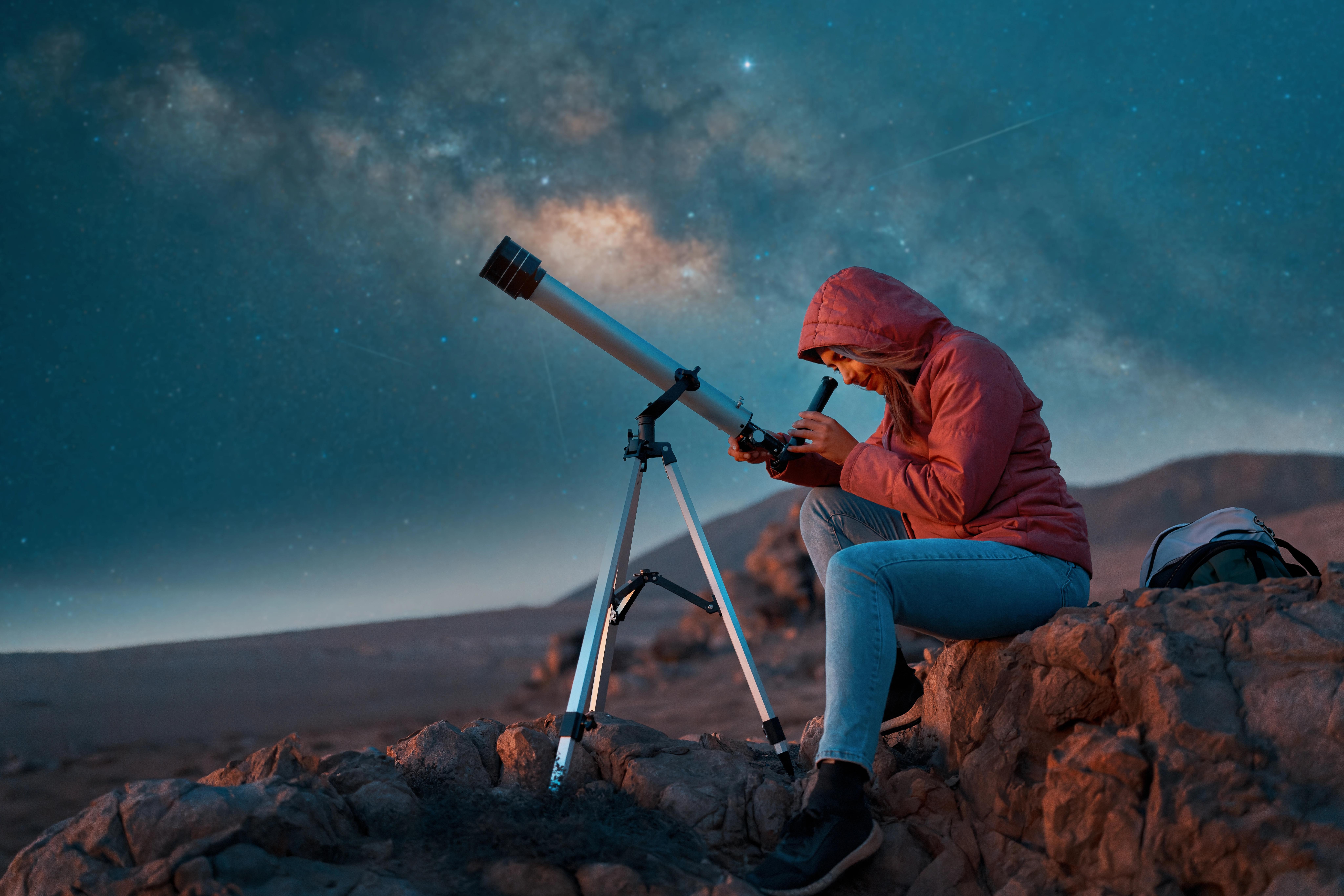 Teple oblečená žena s teleskopem pozorující noční oblohu.