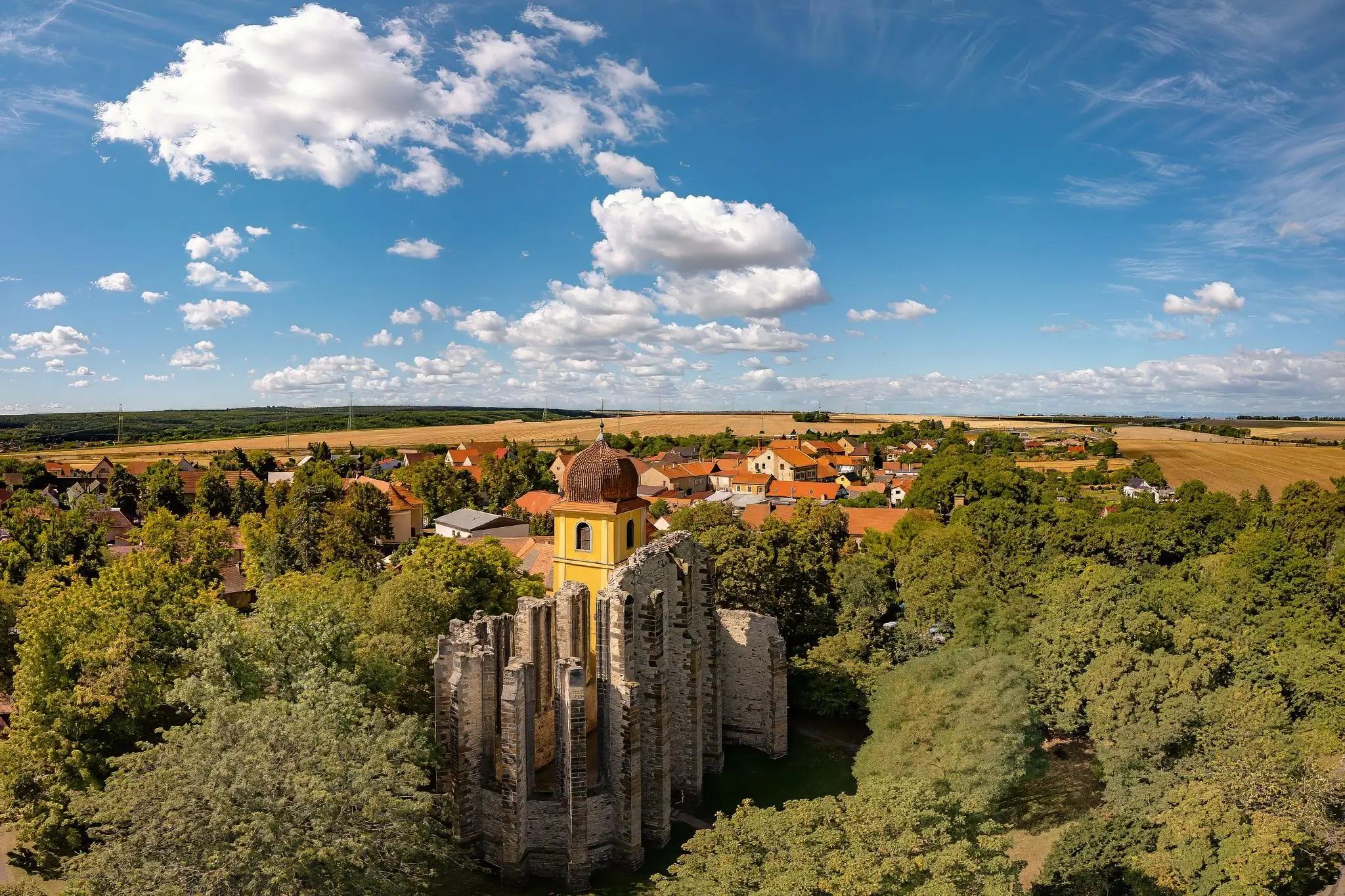 Letecký záběr na nedostavěný chrám Panny Marie v Panenském Týnci. Je obklopený několika zelenými stromy. V pozadí za ním jde vidět menší městys.