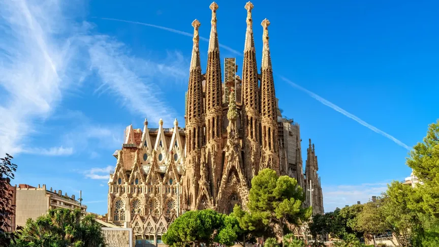 Na obrázku je známý španělský kostel Sagrada Família, tyčící se do modrého nebe. Vidět jdou okolní stromy a nalevo pár okolních budov.