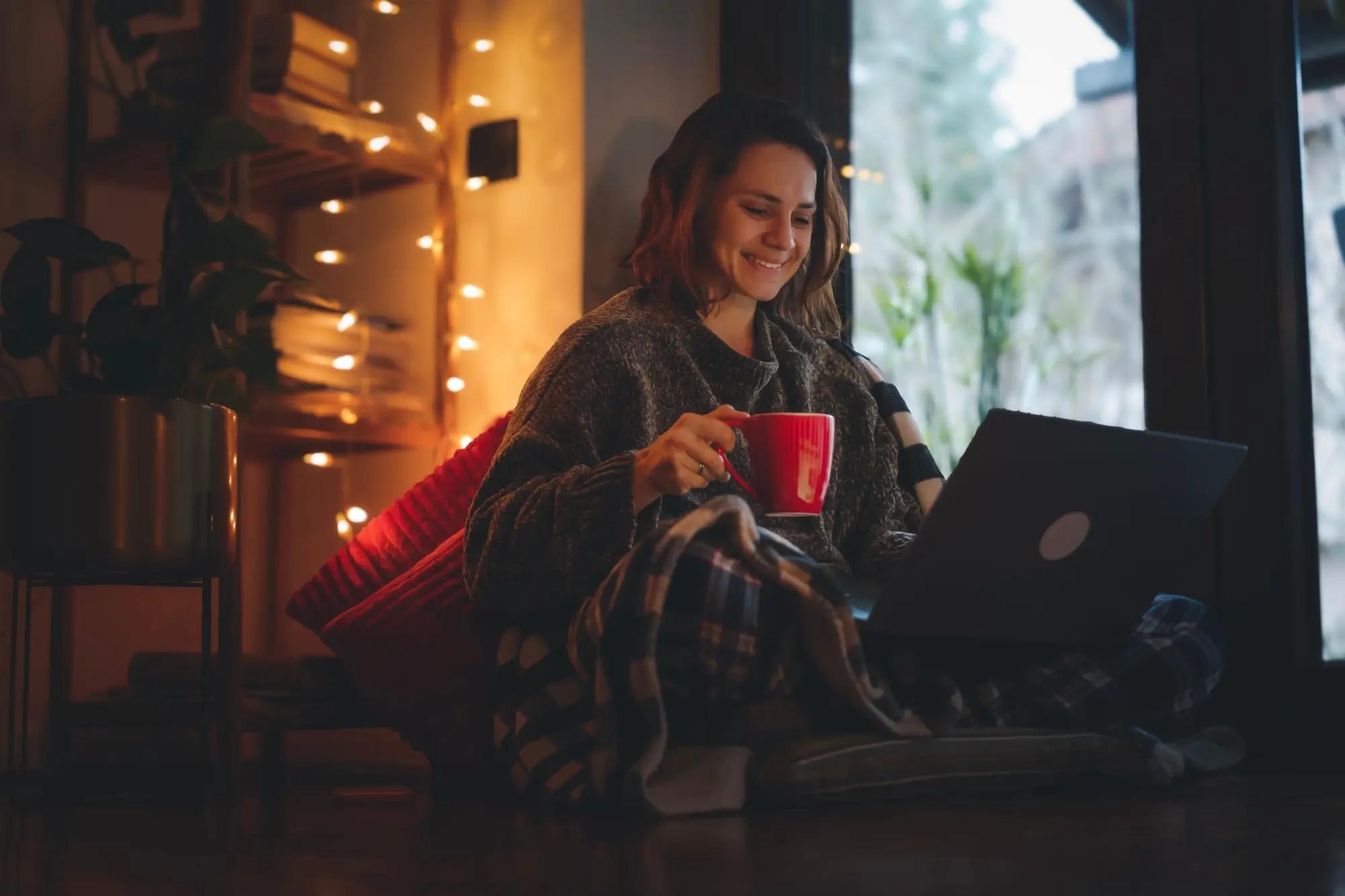 Žena sedí doma v dece, pije čaj a pracuje na počítači