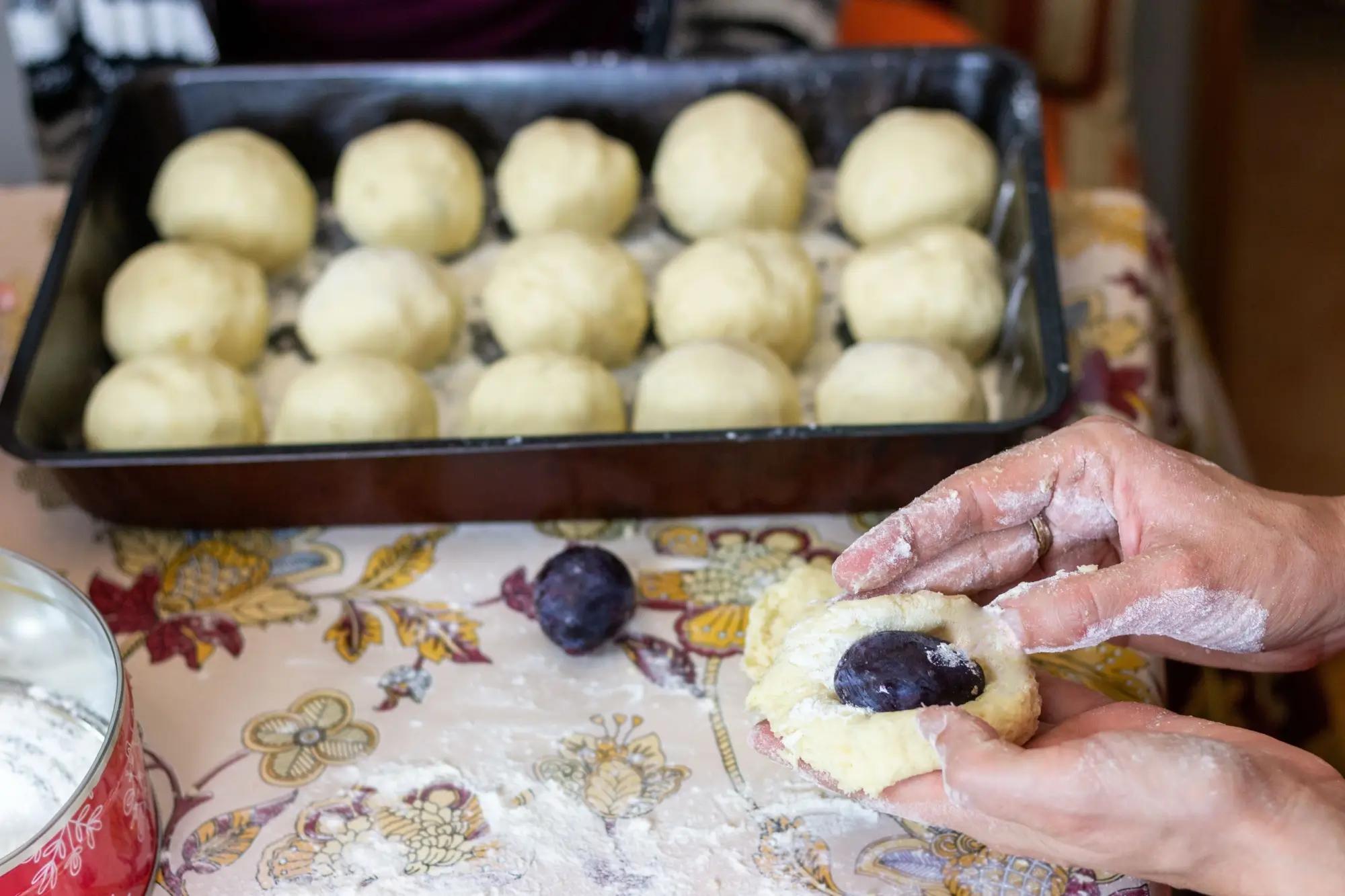 Detail ženských rukou vyrábějící švestkové knedlíky, v pozadí plech z připravenými syrovými knedlíky