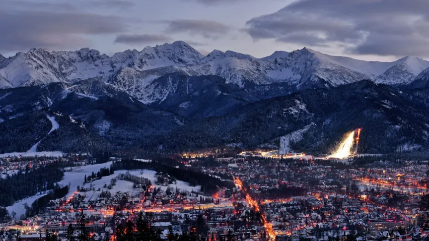Panoramatický obrázek na město Zakopané. Vidět jde město, protože je už trochu více tma, svítí tam světla. Je nasněženo a za městem jdou v dáli vidět také hory.