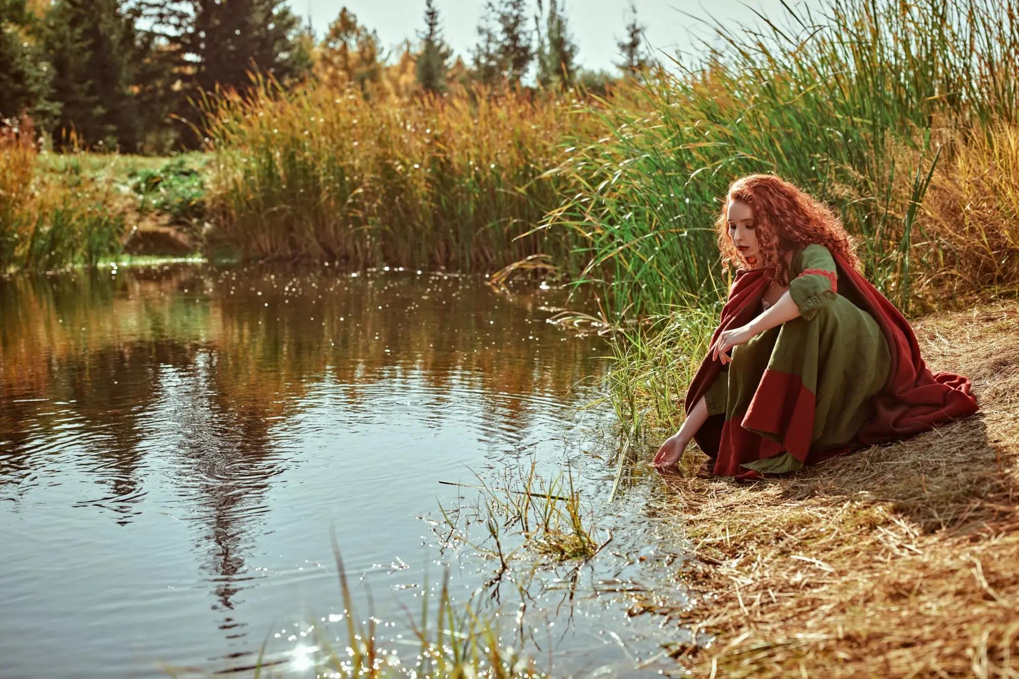 Mladá rusovlasá žena v keltském oděvu sedí u vody