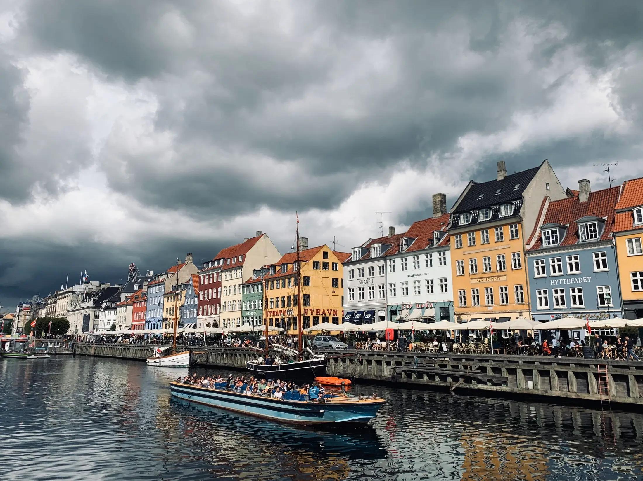 Záběr na hlavní město Dánska, Kodaň. Vidět je kanál s vodou, na kterém pluje loď a na břehu ho lemují barevné domky.