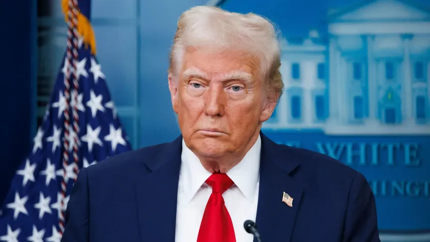 Fotka americkéh prezidenta Donalda Trumpa. Má na sobě modrý oblek s bílou košili a červenou kravatou. Za ním jde vidět logo Bílého domu a americká vlajka. Tváří se neutrálně.