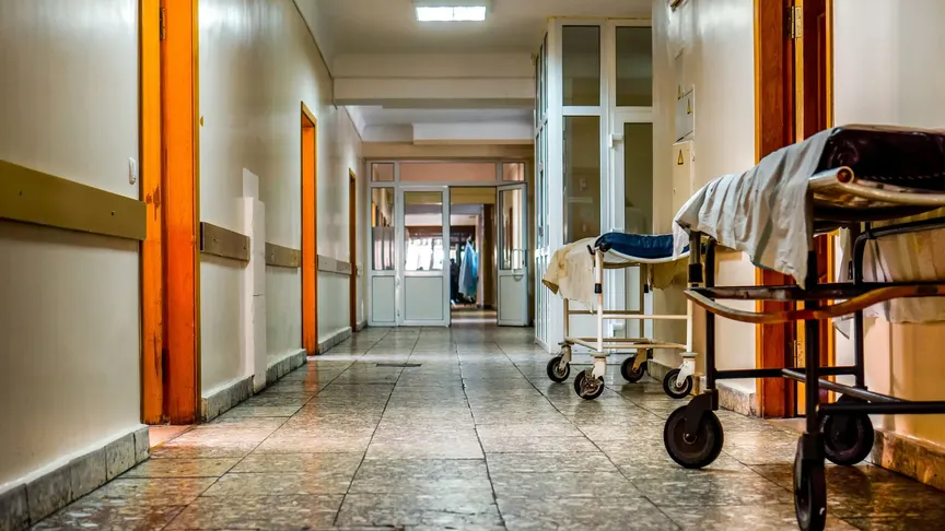 Záběr na chodbu starší nemocnice. Má světlé stěny a lehce popraskané kachličky na podlaze. Nalevo jsou vidět starší lehátka sloužící k převozu pacientů.