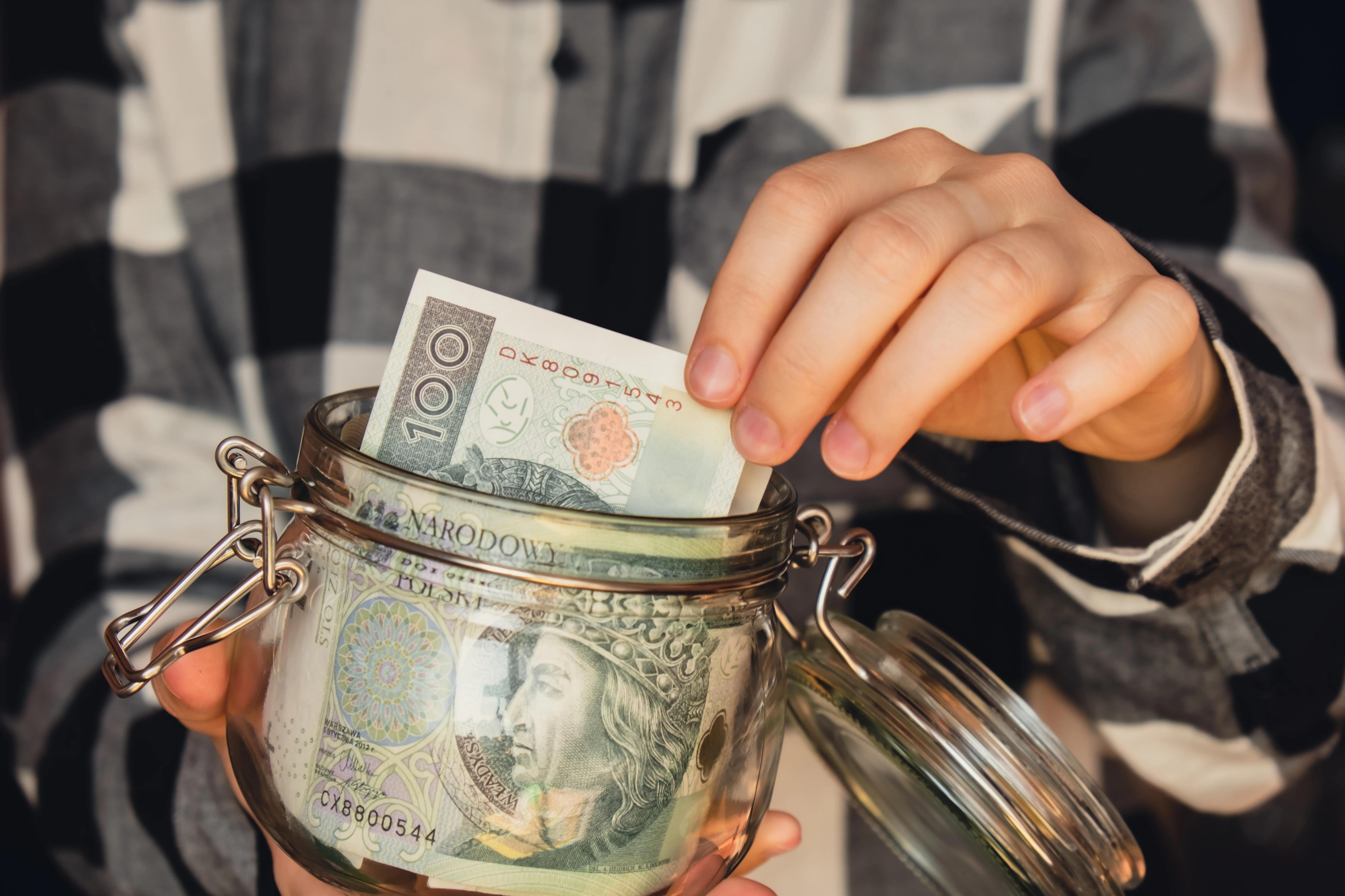 Postava v černobílé kostkované košili drží malou zavírací sklenici, z které vytahuje papírovou bankovku