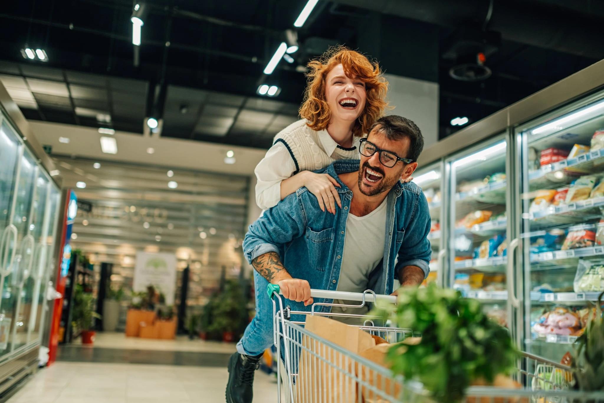 Rozesmáta žena na zádech muže tlačícího nákupní vozík v supermarketu.