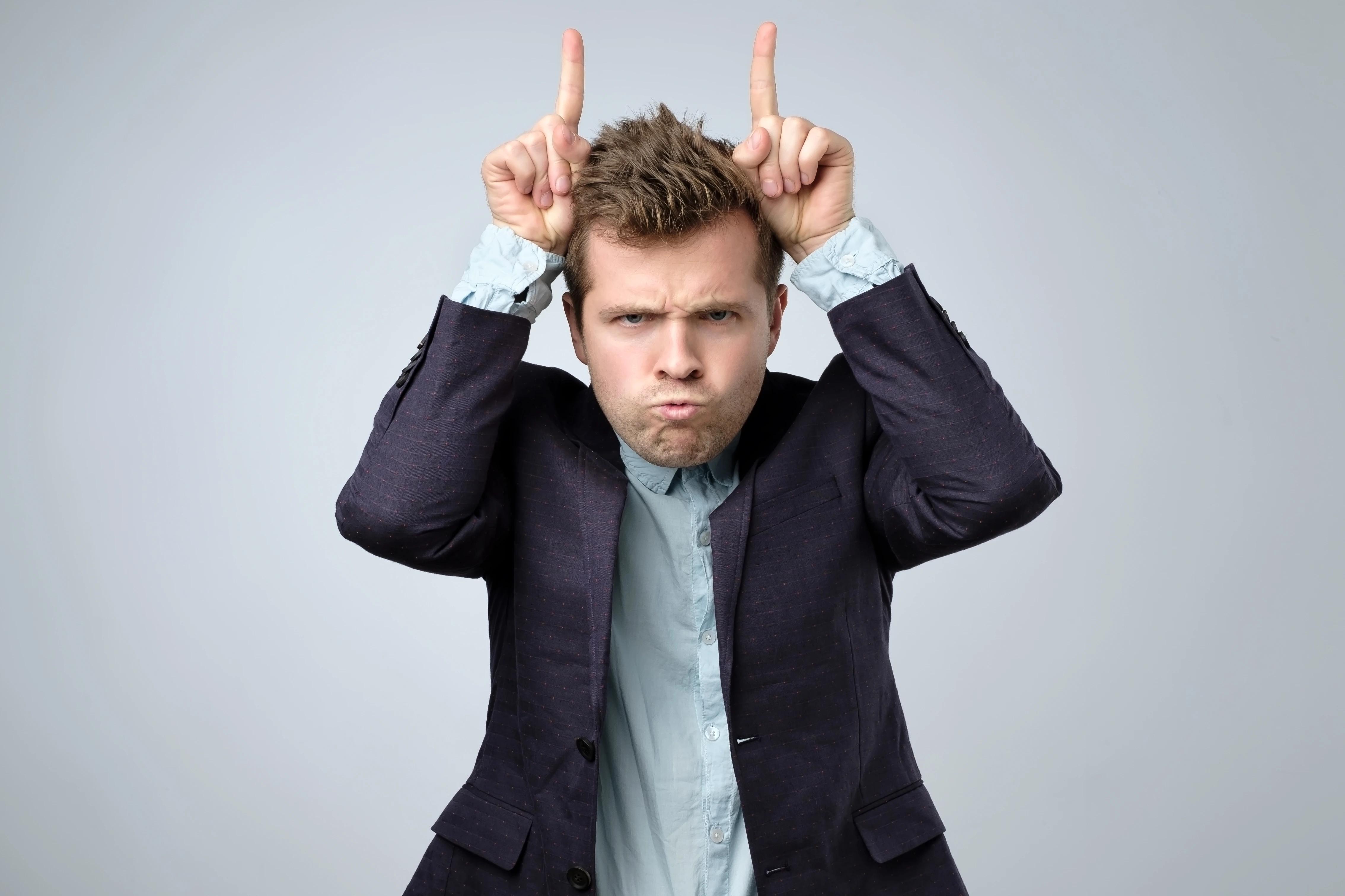 Hravý pohledný vousatý muž v obleku se dívá do kamery s legračním výrazem a prsty ve tvaru kravských rohů