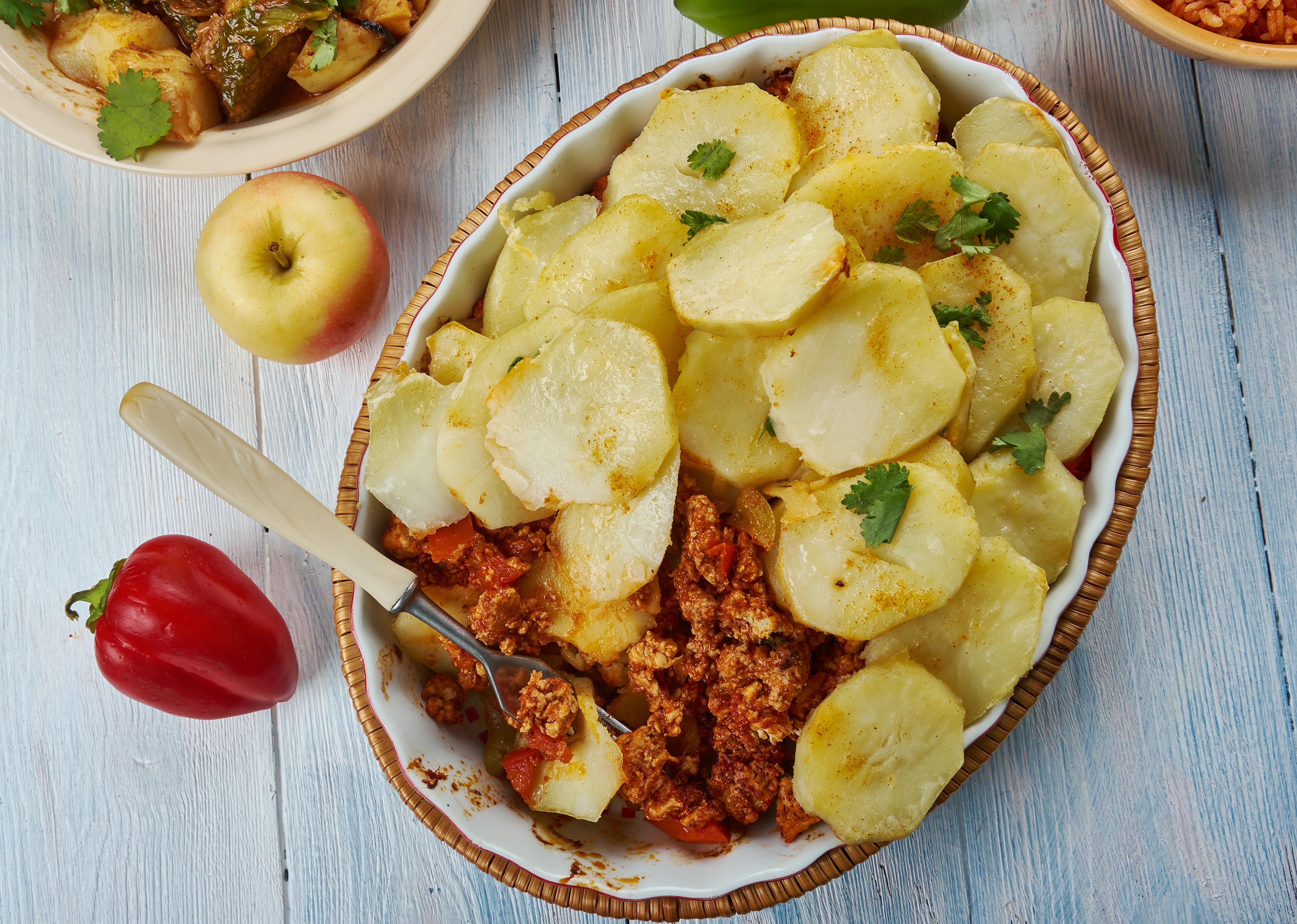 Srbská bramborová musaka na oválném talíři, vedle kterého je červená paprika a žluto-červené jablko