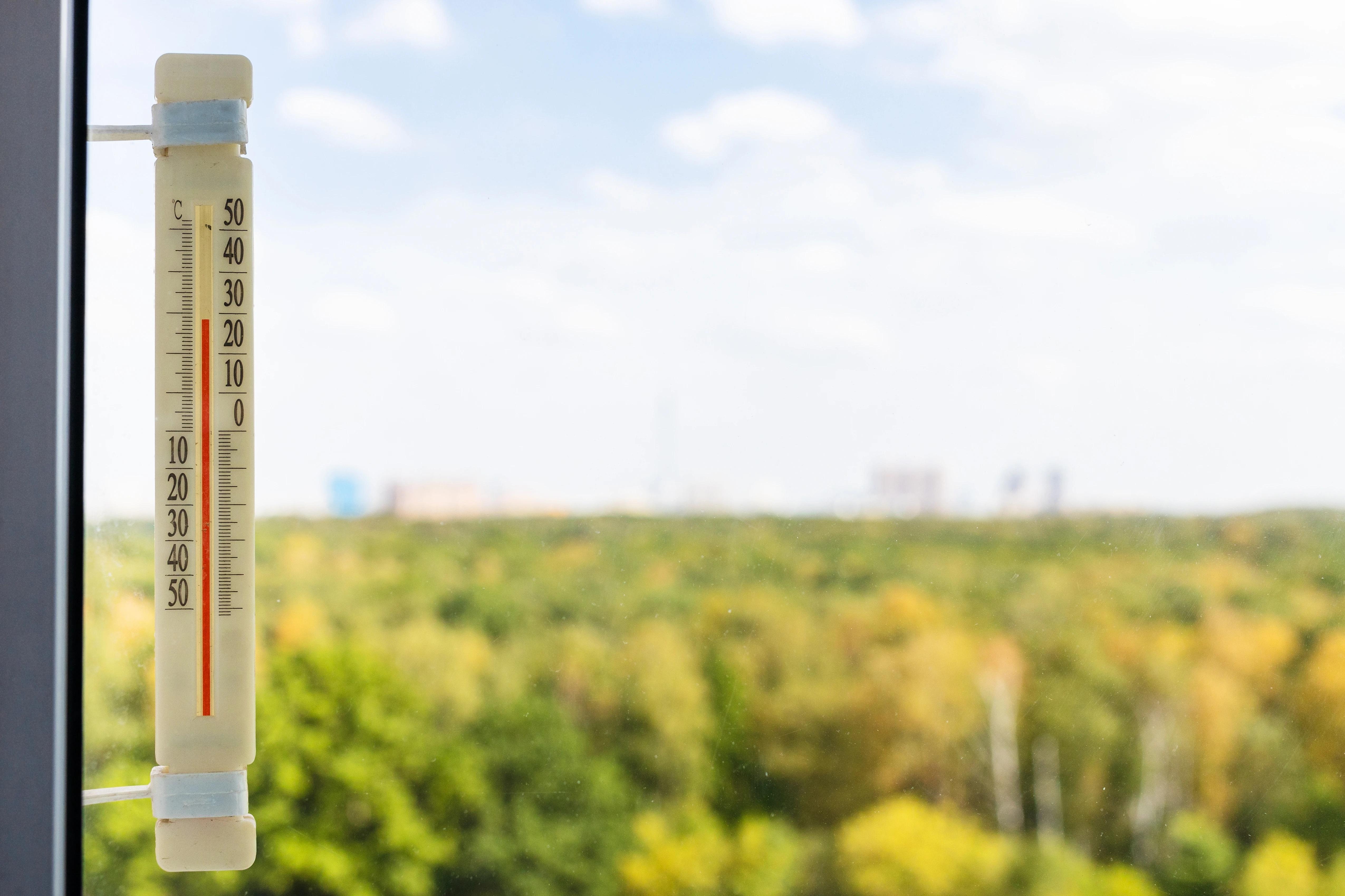 Pohled na teploměr umístěný v okně, za kterým jsou zelené stromy a modrá slunečná obloha s bílými mraky