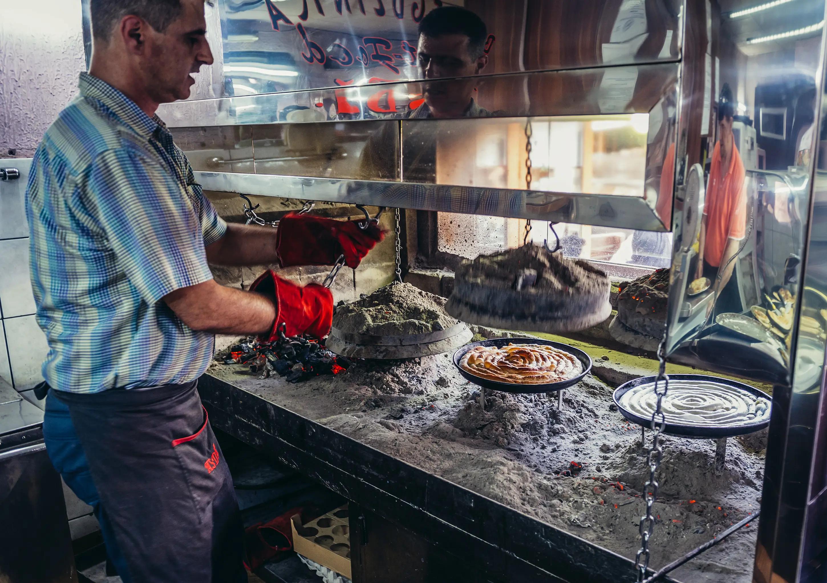 Mladý muž v pruhované modro-bílé košili se zástěrou u pasu připravuje takzvaný rolovaný burek v restaurační kuchyni v Sarajevu