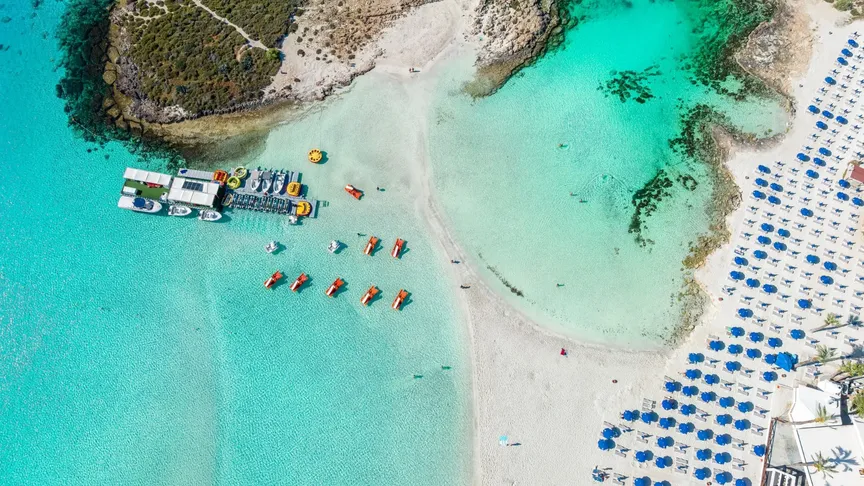 Letecký záběr na pláž Nissi, na které leží lehátka a slunečníky. Písek je skoro bílý, moře krásné tyrkysové.